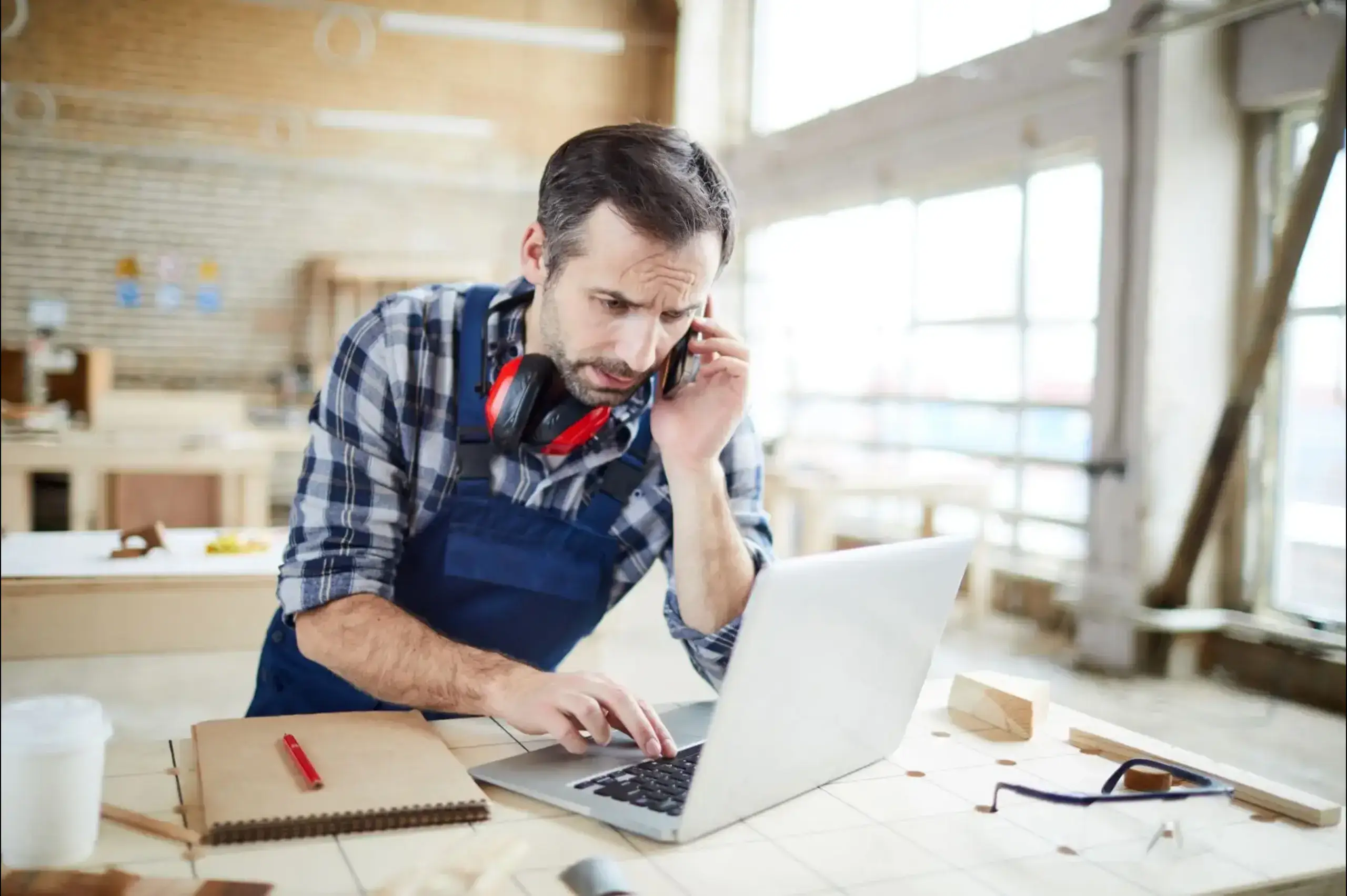 A man leaning on a table, with his right hand checking his laptop and his left calling someone on phone