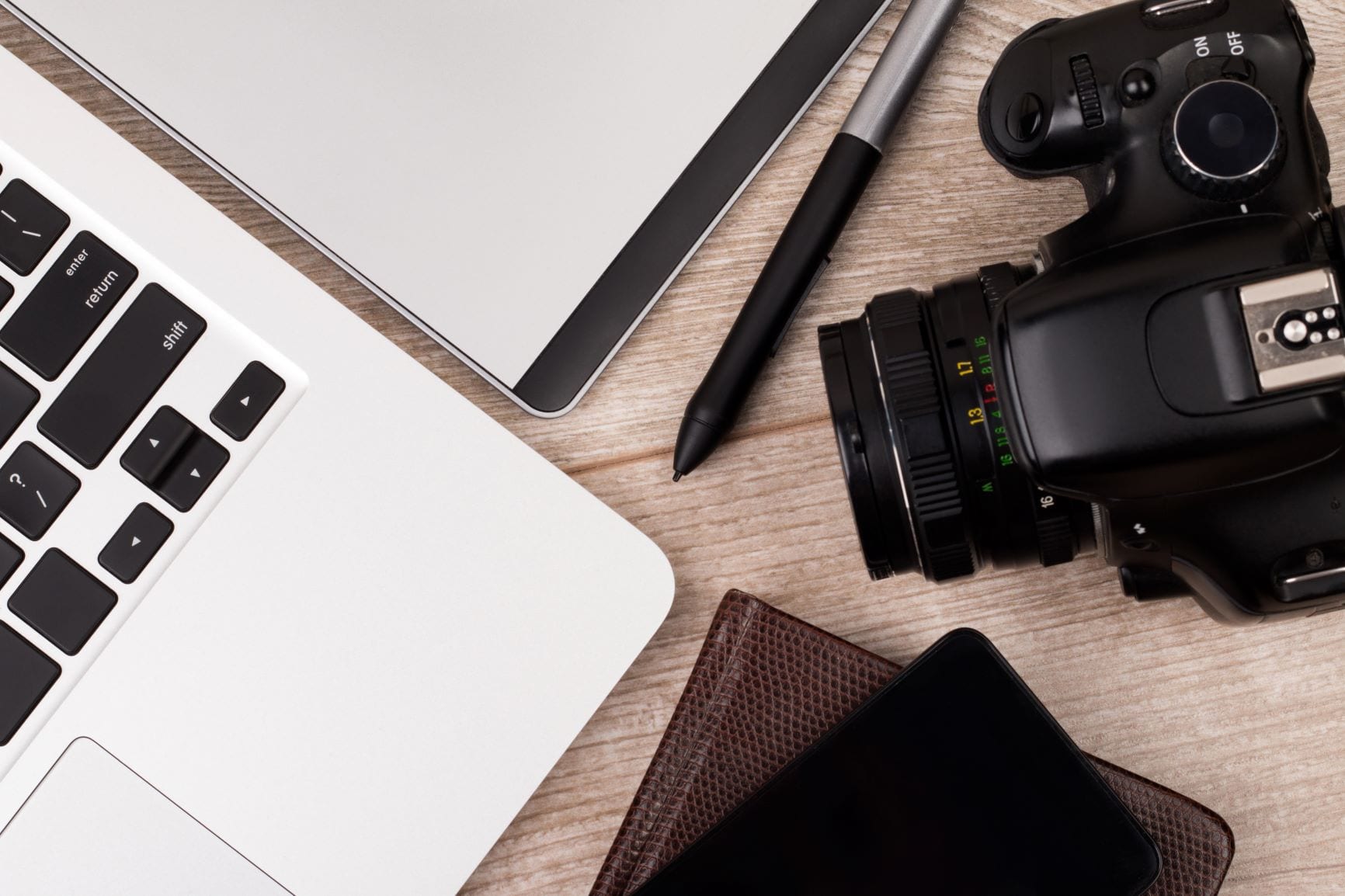 A top view of a photographer or graphic designer's workspace with a laptop, tablet, phone, and camera on a wooden table