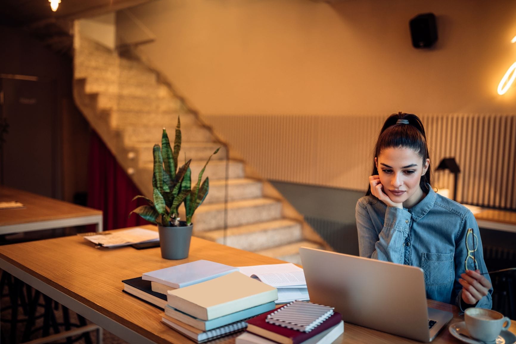 A woman staring intently at her laptop screen with the desk cluttered with notebooks and a cup of coffee on the side