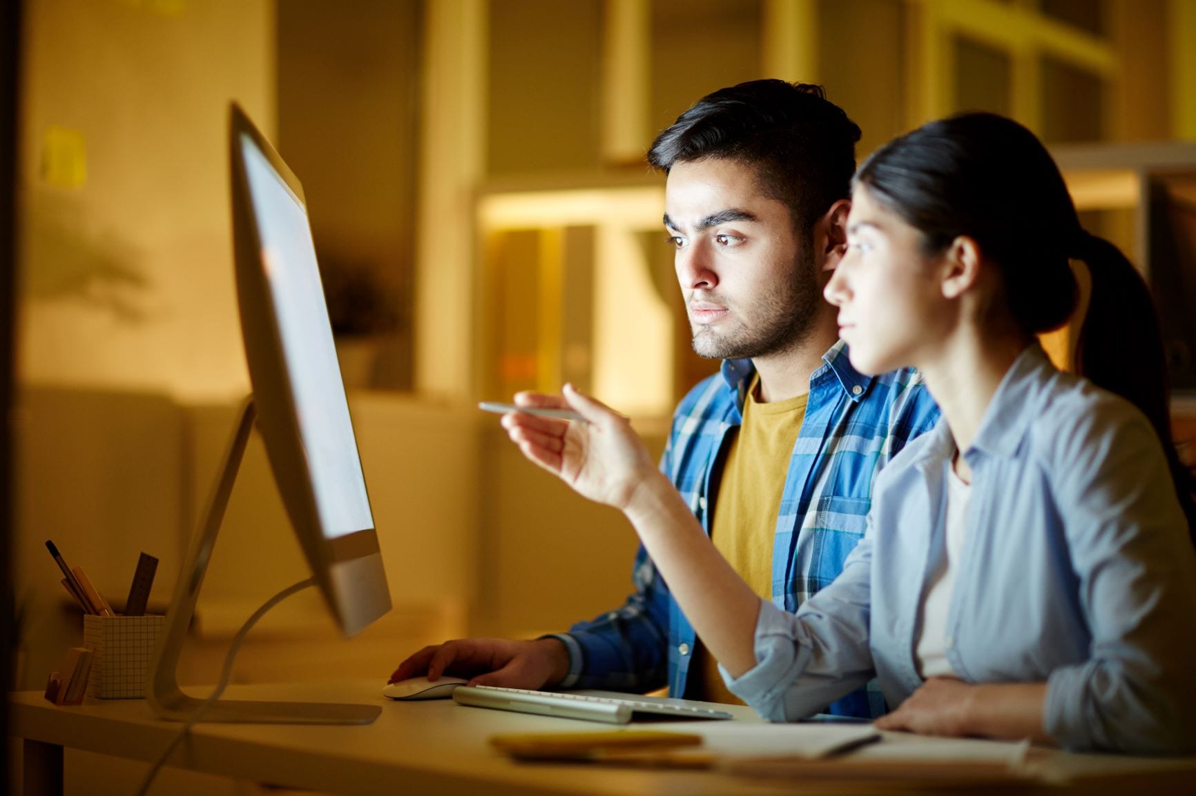 A man and woman sitting in front of a computer, with the woman pointing at the screen while the man looks on intently