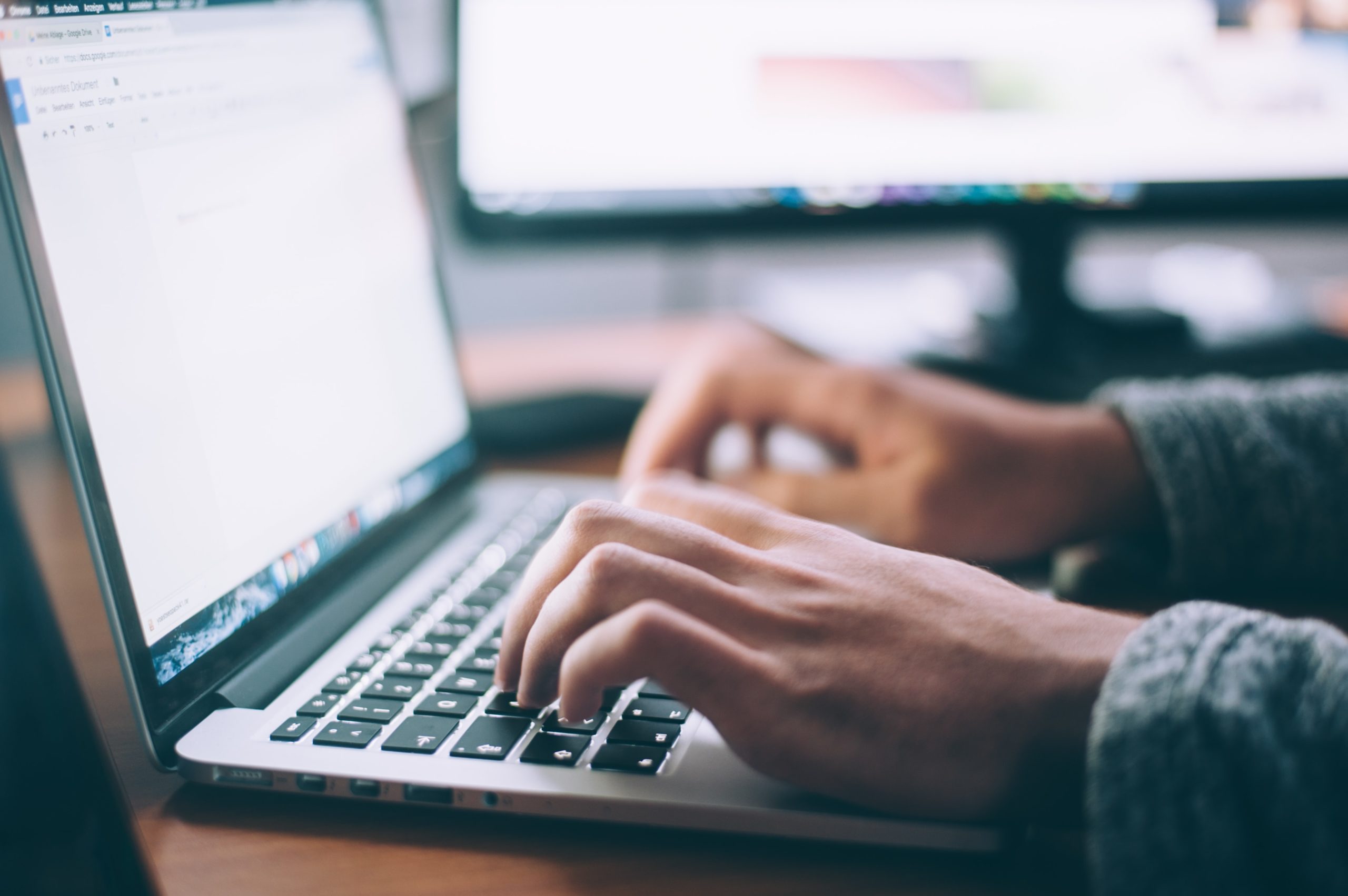 Hands typing on a laptop keyboard placed on a desk with a side computer