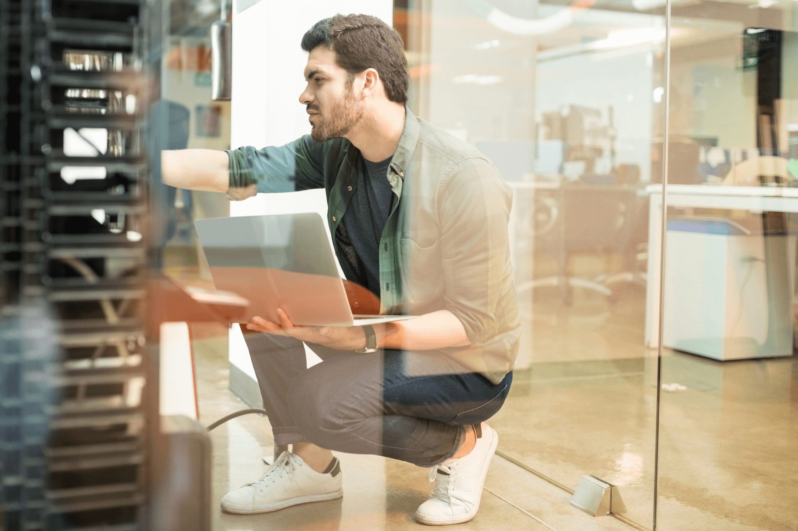 A web designer kneeling and holding a laptop on his right while checking the backup server